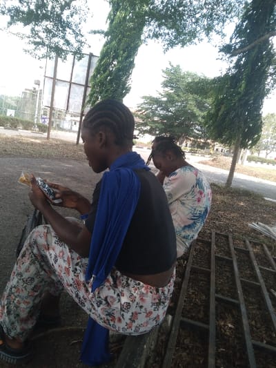 The image depicts an outdoor scene in Osu, Nigeria, featuring two individuals seated on a low ledge under bright daylight. In the foreground, a person with cornrows, wearing a black short-sleeved top, white trousers with a floral pattern, and a blue fabric draped over their left shoulder, holds a smartphone in their left hand and a small, wrapped item in their right, looking downwards. Adjacent and slightly behind them, a second individual, seen from the back, wears a light-colored floral patterned top and also has braided hair, looking downwards. The ground beneath the individuals is covered with dry leaves and dirt. To the right of the seated figures, a metal grid structure lies on the ground. The background features several tall trees, including slender evergreens, and distant urban structures such as a large billboard frame and buildings, visible through the foliage.