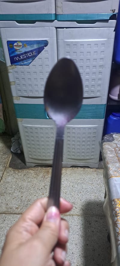 In the foreground, a hand is visible holding a spoon, which is out of focus, with a pinkish tint on the tips of the fingers and a purple hue on the spoon itself. The background consists of two white plastic drawers with textured square patterns. Between the drawers, a turquoise striped design is visible. A blue label with white text, including "MAJESTIQUE" and "OROCAN," is affixed to the lower drawer. The setting appears to be indoors, with a textured floor made of small stones visible to the right of the drawers. A white tablecloth with a gold pattern is draped over something in the upper right portion of the image. The overall lighting suggests an indoor environment, possibly during the day. The scene is casual and domestic.