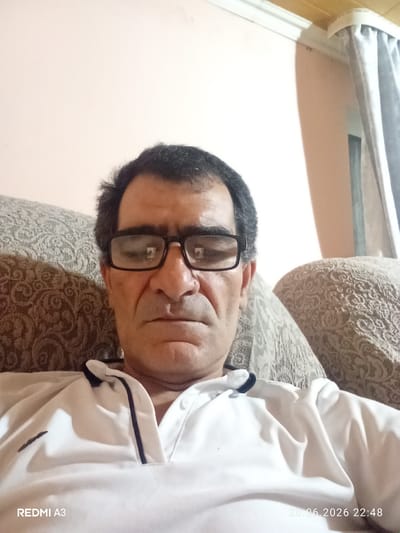 A man wearing glasses and a white polo shirt is pictured from the chest up, looking at the camera. He is seated on a sofa with a patterned fabric. The background shows a wall and a curtain. A timestamp in the lower right corner of the image reads "20.06.2026 22:48".
