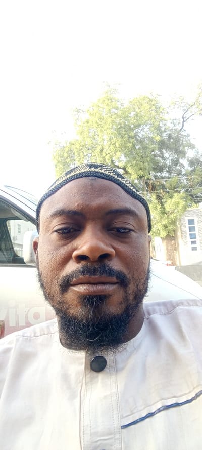 A man wearing a cap and light-colored clothing poses for a selfie outdoors. He has a beard and mustache, with a serious expression. In the background, there's a vehicle with writing on it and a large tree with sparse leaves. A building with windows is also visible behind the tree. The lighting is bright, indicating daytime.