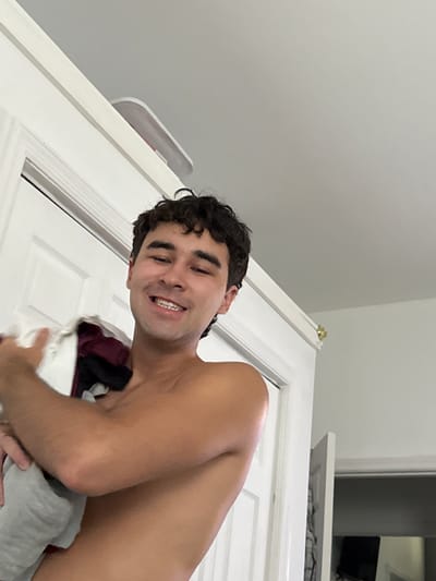 A young man with dark, curly hair smiles and holds a pile of folded laundry to his chest. He is shirtless and his skin is tanned. He appears to be standing in a doorway. The background consists of white doors and a light gray ceiling. On top of the door frame, a white object with a red accent is visible. The scene takes place in a residential interior, likely a home in Bellmore, United States of America.