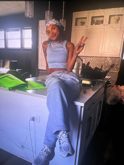 A young woman with a bandana on her head and a light blue crop top is sitting on a kitchen island. She is wearing grey sweatpants and grey sneakers. She is holding up two fingers in a peace sign with her right hand. The kitchen has white cabinets with decorative glass fronts. On the island are two green yoga mats and a black utensil holder filled with kitchen utensils. There is a roll of paper towels to the left of the utensil holder. To the right of the island are plastic bags filled with items. An electrical outlet with a plug in it is visible on the wall below the island. The scene appears to be indoors, in a kitchen. The location is not discernible from the image.