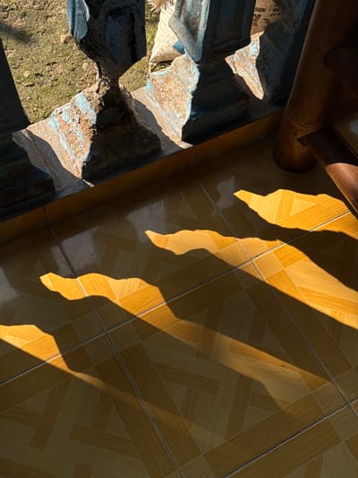 The image captures a sunlit tiled floor with dramatic shadows cast by an overhead structure, likely a balcony or railing. The tiles are a warm, yellowish-brown, arranged in a diagonal pattern. The shadows are sharp and angular, suggesting a low sun angle, possibly late afternoon. The background shows a weathered, blue-painted concrete railing with visible rust and rough texture, alongside dry earth and some vegetation. A portion of a wooden structure, possibly a table or chair leg, is visible on the right side. The scene appears to be an outdoor space, possibly a patio or walkway, in Kalirancang, Indonesia, characterized by the interplay of sunlight and shadow on the textured surfaces. There are no people or animals visible in the frame.