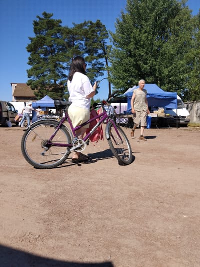 A medium shot captures a woman from behind, holding a smartphone, standing next to a purple bicycle. She is wearing a white t-shirt and a light-colored skirt. The bicycle has a pink bag attached to its rear. In the background, a man with gray hair and wearing a tan sleeveless shirt and shorts walks away from a market stall with blue tents. Behind him, a white van is parked. The scene is set outdoors on a dirt surface under a clear blue sky, with large trees visible in the background. The location is a market in Irshansk, Ukraine.