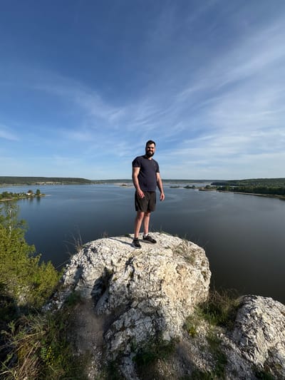 A man stands on a rocky precipice overlooking a body of water. He is wearing a dark T-shirt and shorts, facing the water with his left arm extended. The sky is blue with scattered clouds. The opposite bank of the water features a line of trees and buildings. The location is Gorbunyata, Russia.