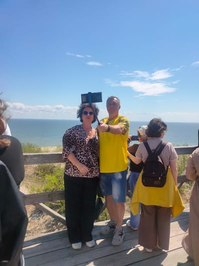 In Morskoye, Russia, a group of people are standing on a wooden platform overlooking the sea. A woman in a leopard-print top and black pants, wearing sunglasses, holds a selfie stick with a phone attached, taking a photo of herself and a man in a yellow t-shirt and denim shorts. To the right of the couple, a woman with a backpack and a yellow shawl is looking away, and another person in a beige dress is partially visible. The sky is blue with scattered clouds, and the sea stretches to the horizon. Green vegetation and sand dunes are visible below the platform.