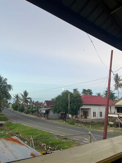 The image depicts a street scene in Turumame, Indonesia, during what appears to be early morning or late evening due to the soft lighting. The foreground is dominated by a corrugated metal roof, likely from a building. A paved road runs horizontally across the mid-ground, with a grassy verge on either side.

Along the right side of the road, a cluster of houses is visible. One prominent house features a white facade and a red tiled roof. Adjacent to it are other buildings, some with wooden or corrugated metal exteriors, and small shops or stalls. Tropical vegetation, including palm trees, is interspersed throughout the scene, suggesting a warm climate.

A single street light on the left side of the road is illuminated, casting a soft glow. There are utility poles with numerous wires crisscrossing the sky. A utility pole with a reddish-brown pole stands out on the right. In the distance, more houses and palm trees line the road as it curves away. A small yellow trailer is parked near one of the buildings. A motorcycle is parked to the left of one of the houses. The sky is a pale blue with a hint of haze. The overall impression is of a quiet, rural or semi-rural street in Indonesia.