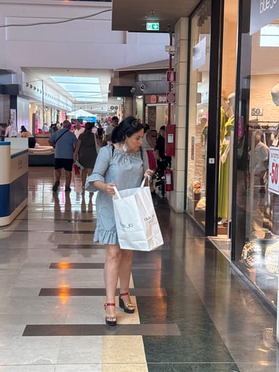 The media file shows a bustling indoor shopping mall in Carini, Italy. Several people are visible, walking and browsing. The main subject is a woman in a striped dress holding a white shopping bag with "blue jo" logo visible. She is wearing heeled sandals. Other shoppers can be seen in the background, some carrying shopping bags. The mall appears to be well-lit, suggesting daytime. The floor is tiled with a pattern of dark and light rectangular tiles, with inlaid lights reflecting on the surface. Signs are visible in the background, including one with "50%". The overall atmosphere is that of a busy retail environment.