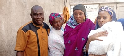 The image displays four individuals standing outdoors in the city of Dan Maliki, Nigeria, positioned against a plain, light-colored wall. From left to right:
1. A bald male, appearing middle-aged, with a dark complexion, wearing an orange short-sleeved collared shirt with a black front panel. He exhibits a slight smile and looks towards the viewer.
2. A young adult female, dark-complexioned, wearing a white head covering and a multi-colored patterned headwrap. Her expression is neutral, and she gazes slightly to her left.
3. A middle-aged female, dark-complexioned, wearing a dark head covering and a magenta-colored wrap adorned with light blue patterns. She wears gold earrings and maintains a neutral gaze towards the camera.
4. A young adult female, dark-complexioned, wearing a blue patterned headscarf and a nose piercing on her right nostril. She holds a white, bundled, fluffy object, consistent with an infant wrapped in a blanket, against her chest. She looks directly at the camera.
The background shows minimal structural details, including a faint outline of a doorway or aperture and a small, dark object hanging on the wall behind the central figures. The lighting suggests daytime.