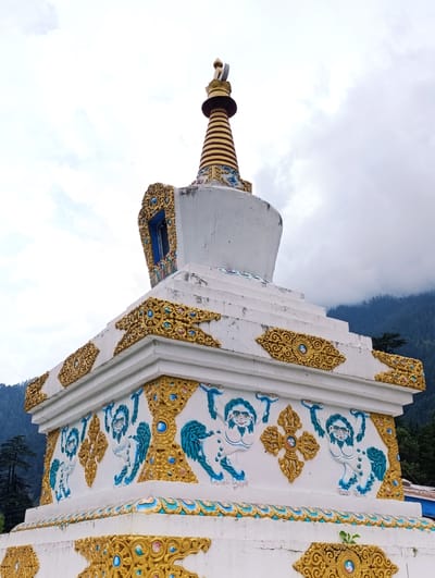 A Buddhist stupa, a white structure with gold and blue ornamentation, stands in Gadherni, India. The stupa features carved figures of mythical creatures and intricate decorative patterns in gold. A multi-tiered conical finial in gold and brown crowns the stupa, topped with a small gold spire. The structure is set against a backdrop of misty, forested mountains under a cloudy sky. The lower portion of the stupa is adorned with relief carvings of white, lion-like creatures with blue manes and details. The stonework is detailed with circular motifs and floral designs, predominantly in gold. The overall impression is of a serene, sacred site integrated with the natural landscape.
