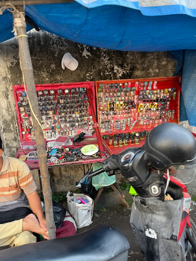 A street vendor is displaying a variety of watches for sale at a market stall in Gedang, Indonesia. The stall is set up under a blue tarp, with a concrete wall as the backdrop. The watches are displayed in two red-backed cases, one on top of the other, and there are also watches and accessories laid out on a table in front of the cases. A man, presumably the vendor, is sitting on the left side of the image, wearing a striped shirt and light-colored pants. A motorcycle is parked in front of the stall, with a black helmet resting on the handlebars. There are also several bags and containers scattered around the stall. The ground is wet, suggesting it may have recently rained.