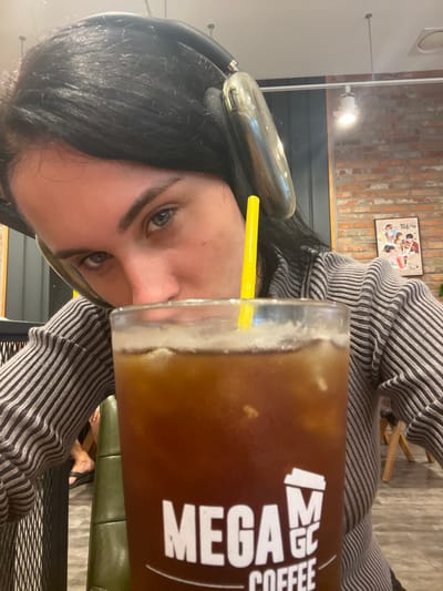An individual, likely female, with dark hair and light eyes, is depicted from a low angle within a MEGA MGC COFFEE branch in Gangseo-gu, South Korea. The person wears large, reflective silver over-ear headphones and a grey and black striped long-sleeved shirt. Their face is partially obscured by a large, clear glass of iced dark brown coffee with a yellow straw, bearing the white "MEGA MGC COFFEE" logo. The individual's eyes are directed at the viewer. The background features an interior cafe setting with a brick accent wall on the right, on which a framed poster with multiple figures and Korean text, including "청춘기록" and "BTS," is mounted. Ceiling spotlights illuminate the space. Elements of cafe furniture, including a green upholstered seat and dark metal grating, are partially visible. A foot wearing a black flip-flop belonging to another individual is visible in the lower left background.