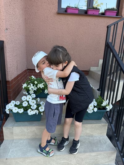 Two children, a boy wearing a white cap and a white t-shirt with a graphic print and blue shorts, and a girl wearing a white t-shirt and a black vest with black shorts, are hugging on a set of concrete stairs in front of a textured pink wall. The boy is kissing the girl on the cheek. White petunias are planted in green rectangular planters on the stairs. A black metal railing is visible to the right. The background shows a window with three purple planters containing plants. The image appears to be taken outdoors on a sunny day. No specific identifiable landmarks or indications place this scene in Kolomyia, Ukraine.