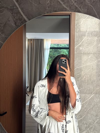 A person is taking a mirror selfie in a modern bathroom. The person, with long dark hair, is wearing a black bralette and a white robe with black lettering. They are holding a smartphone in their right hand, obscuring their face. The bathroom features a large, oval mirror framed by wood and marble. In the reflection, a window with sheer curtains reveals lush green trees outside, suggesting a location within a natural setting, possibly in the city of Bakovići, Montenegro. The walls are adorned with a grey marble-like finish. A wooden door with a black handle is visible to the left. The floor appears to be made of wood.