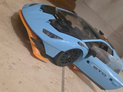 The image shows a close-up, angled view of a blue toy car, possibly a Lamborghini, on a tiled floor. The car has orange accents on its lower body and spoiler. The word "STO" is visible on the side door. The floor tiles appear to be light brown with darker grout lines. The background is out of focus and shows a blurred brown surface, possibly a table or furniture. The lighting is dim and appears to come from the top right of the image. There are no people or animals present. The overall scene suggests an indoor setting, likely a home, and the car appears to be stationary. There is no indication of the car's location in Ladispoli, Italy, beyond the provided prompt.