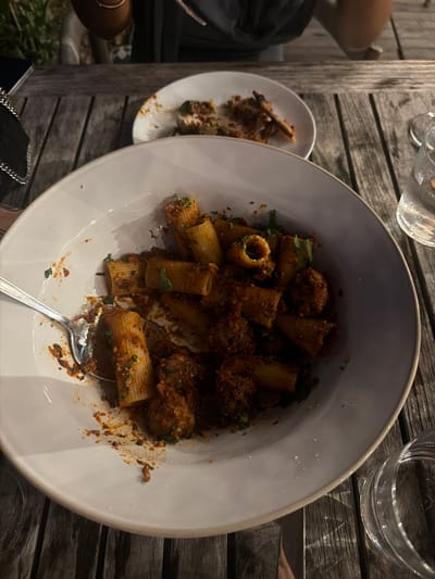 A close-up, top-down view shows a white ceramic bowl filled with rigatoni pasta and meatballs in a red sauce, resting on a weathered wooden table. A silver fork is partially submerged in the pasta. In the background, out of focus, a second white plate holds a small portion of food. A glass of water is visible to the right of the main bowl. The scene appears to be outdoors, possibly a restaurant patio, given the wooden table and hint of greenery in the upper left corner. The lighting suggests it is evening or dusk. The location is identified as Montauk, United States of America.