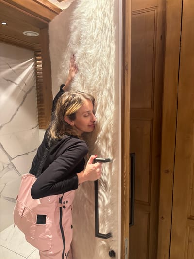 A female individual is observed indoors in St. Christoph, Austria. She is depicted with blonde hair, partially braided, and wearing a black long-sleeved top over pink ski bib overalls. Her eyes are closed as her head rests against a door covered in white, long-pile faux fur. Her right hand, with red nail polish, is placed gently on the fur-covered surface. Her left hand grips a dark, L-shaped door handle affixed to the same fur-covered door. To the right of the fur-covered door, a light-colored wooden door is ajar, revealing an interior wooden surface beyond. The immediate environment includes a wall tiled with white marble featuring grey veining on the left, and light-colored wooden paneling on the right. A wooden ceiling or soffit containing a recessed light fixture is visible in the upper left quadrant.