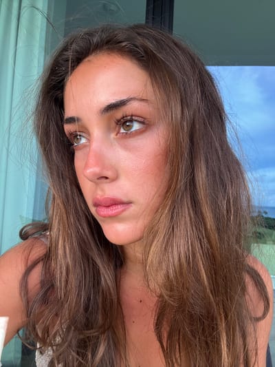 A young woman with light brown hair and olive skin gazes at the camera. Her hair is wavy and long, framing her face. Her eyes are almond-shaped, and her lips are full and glossy. She appears to be outdoors, with hints of sky and buildings in the background. The overall composition suggests a casual, natural snapshot.