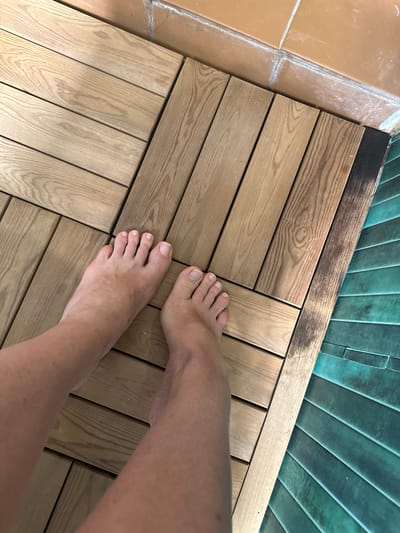 The image shows a person's bare feet on a wooden deck. The deck is made of light-colored wooden planks arranged in a herringbone pattern. To the right of the deck is a dark strip, possibly a drain or edging, and beyond that, a surface of teal-colored, ribbed tiles. In the upper right corner of the image, there are terracotta-colored tiles with white grout. The scene appears to be outdoors, possibly a patio or poolside area. The location is not specified in the image and therefore cannot be identified as Novoalexandrovka, Russia.