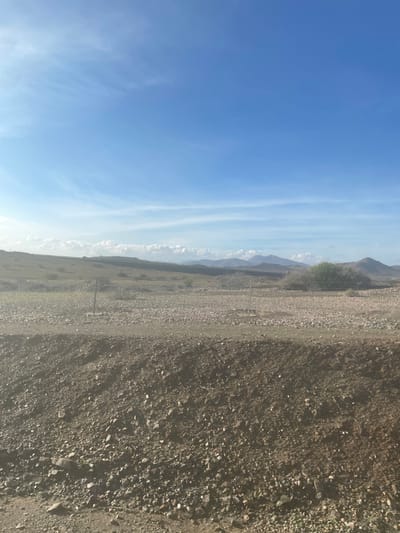 The image depicts a wide, vertically oriented landscape. The left half of the frame is dominated by a clear, bright blue sky with sparse, wispy white clouds stretched horizontally across it.
A horizontal band across the middle of the image separates the sky from the foreground. This band showcases a distant, hazy mountain range or hills, appearing light greyish-blue against the sky. Below these mountains, in the mid-ground, is a flatter expanse of dry, yellowish-brown terrain with scattered, low-lying vegetation. Two slender, dark vertical posts, possibly fence posts or markers, are visible in this mid-ground area, one roughly central and another further right.
The right half of the image consists of rocky, gravelly ground, appearing in shades of dark brown and grey. This foreground area is covered with numerous small stones and pebbles, suggesting a rough, arid surface, possibly part of a dirt road or track.
The scene captures a static natural environment under bright daylight, indicating a clear and sunny day. There are no people, animals, or discernible text in the image. The overall impression is one of vastness and natural desolation, consistent with an arid or semi-arid region such as Jbilate, Morocco.