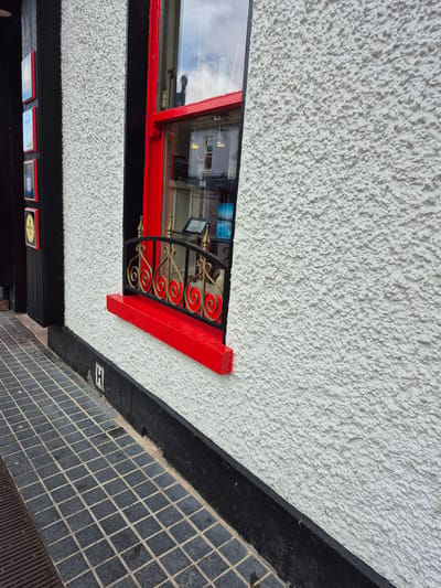 The image depicts the exterior of a building in Carrick-on-Shannon, Ireland. A white, rough-textured wall with a bright red window frame is a prominent feature. The window has a decorative black metal grate with gold accents in front of it. Below the window, a red windowsill protrudes. To the left of the wall, a black vertical structure with several framed square signs, some with red borders, is visible. The ground in front of the building is paved with dark grey square tiles, and a metal grate is partially visible on the left. A black plinth runs along the base of the white wall.