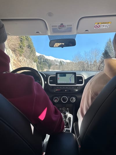 The image depicts the interior of a vehicle in Gradačka Poljana, Montenegro, viewed from the rear passenger seat, looking forward through the windshield. The driver, wearing a dark red long-sleeved top and grey headwear, is positioned on the left with a left hand on the steering wheel. A front passenger, wearing a light beige long-sleeved top, is visible on the right. The dashboard features a central infotainment screen displaying a navigation map with a route indicating "38 km" distance and a blue path. Below the screen are climate controls and a manual gear shifter. Sunlight casts bright reflections on the driver's left arm and the central console. Through the windshield, a clear blue sky is visible above snow-capped mountains in the distance. The immediate environment outside shows a winding road flanked by sparse trees and a rocky embankment to the left. Evergreen trees are also present, particularly on the right side. The car's rearview mirror is partially obscured by a blue sunshade. An overhead console contains interior lights and a red SOS button.