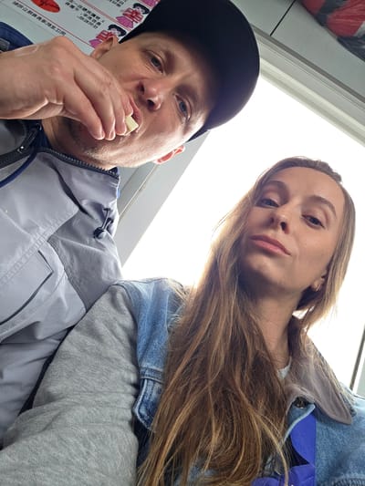 This low-angle selfie captures two individuals, a man and a woman, looking towards the camera against a bright white background, likely an overhead light source or window.
On the left, a man is seen from the chest up, wearing a black baseball cap and a grey and blue zip-up jacket. He has light stubble and blue eyes. He holds a small, light-colored piece of food (resembling cheese or a biscuit) between his thumb and index finger, bringing it towards his mouth as if about to eat. Adjacent to him, on the far left, a white panel displays red and black text, clearly identifiable as Chinese characters, alongside stylized, cartoon-like illustrations of people, possibly women in uniform or traditional attire.
On the right, a woman is visible from the chest up, her long, light brown hair flowing over her shoulder. She wears a light blue denim jacket over a grey long-sleeved top. She looks directly at the camera with a calm and neutral expression, her face slightly angled.
The bright background and the indoor-like setting suggest the image was taken inside a vehicle or building. The Chinese text on the wall serves as a significant location cue. The overall atmosphere is casual, capturing a shared, candid moment.