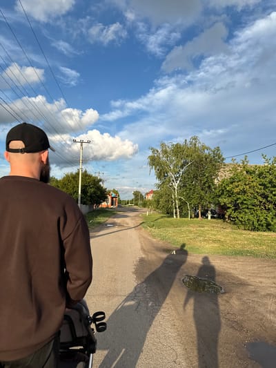 The image captures a street scene in Rossosh, Russia, taken from behind a person wearing a brown sweatshirt and a black baseball cap. The person is pushing a stroller, the top of which is visible. The street is unpaved, with cracks and patches of asphalt, and stretches into the distance, lined by trees and buildings. Power lines are strung overhead. Two shadows are cast on the street, one of which is pointing in the direction the person is walking, and the other is longer and more indistinct. A puddle of water reflects the sky on the road. The sky is blue with scattered white clouds, suggesting daytime, likely late afternoon due to the angle of the shadows and the warm light. A few houses and trees are visible on either side of the street. The overall atmosphere is calm and rural.