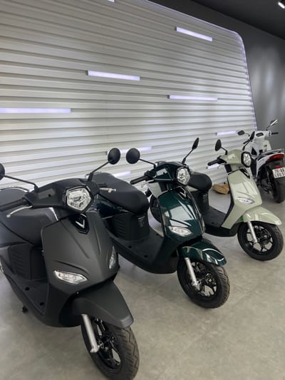 The image displays an interior view of a showroom featuring four modern scooters, with no individuals present. In the foreground left, a matte black scooter is visible, characterized by a V-shaped emblem on its front fairing, a black seat, and circular rearview mirrors. To its right, a dark green scooter of similar design is displayed. Further right, a light beige scooter, also consistent in design with the preceding two, is visible. In the background, a white scooter is partially visible, showing its rear fender with a Vietnamese license plate "76-AA 490.5X". The background wall consists of light grey horizontal ridged paneling, interspersed with several horizontal LED light strips emitting white illumination. The floor is covered with light grey square tiles. A small, light-brown cardboard box is partially visible behind the light beige scooter. The "76-AA" prefix on the license plate indicates registration in Quảng Ngãi province, Vietnam. The specific location or address within Vietnam, including Kon Tum as requested, is not discernible from the image.