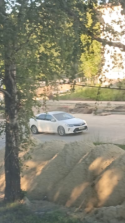 The image shows a white sedan driving on a paved road, partially obscured by trees in the foreground. The car appears to be a Toyota Camry. It is daytime, as indicated by the bright sunlight casting shadows. The scene appears to be an urban or suburban environment, possibly undergoing construction, with piles of sand visible in the foreground and background. There are also buildings and greenery in the distance. The weather seems to be clear and sunny. No people are clearly visible in the image, although a small, indistinct figure is present near the base of a tree in the lower left corner. Some text is visible on the license plate of the car, but it is too blurry to be legible. The overall impression is a snapshot of everyday life in a Russian town.