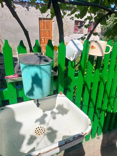 In a rustic outdoor setting, likely in Salhany, Ukraine, an old white enamel sink is prominently featured. The sink, showing signs of age with rust around its rim, is positioned in front of a bright green wooden fence. Above the sink, a turquoise bucket with a black lid hangs, possibly serving as a makeshift water dispenser. A metal scrubber and a pink clothespin are attached to a wooden shelf supporting the bucket.

On the fence, two enamel mugs are hung. One is white with visible text that appears to be a series of numbers and letters. The other mug is cream-colored and decorated with a small strawberry illustration.

The scene is bathed in sunlight, suggesting it is daytime. Shadows from leaves and branches are cast across the sink and fence, indicating the presence of nearby trees or vines. In the background, a weathered building with a wooden door and windows with reddish curtains can be seen. The overall impression is one of a simple, functional outdoor space, possibly in a rural or semi-rural area. The presence of the sink and water bucket suggests an area where washing or cleaning might take place, common in older Ukrainian homes or summer kitchens.