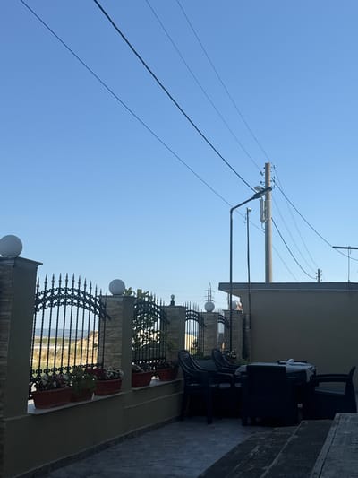 The image depicts an outdoor scene with a clear blue sky and power lines stretching across it. In the foreground, a decorative black metal fence with pointed finials encloses a paved area. Behind the fence, there are several potted plants and some ornamental lights. To the right, a dark-colored outdoor furniture set, including chairs and a table, is visible under an overhang, with a patterned cloth draped over the table. The furniture is placed on a paved patio.
In the background, a body of water, likely the sea, can be seen beyond the fence. The overall setting suggests a residential or hospitality area, possibly a hotel terrace or a private balcony overlooking the water. The time of day appears to be daytime, given the bright sky. The weather seems clear and sunny. There are no people visible in the image. The image is oriented vertically, with the sky occupying the upper portion and the fence and furniture in the lower portion. The location is identified as Slanchevo, Bulgaria.