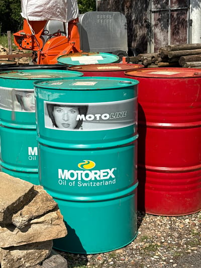 The image shows a stack of oil drums in an outdoor setting. At least three turquoise oil drums are stacked vertically. The label on the barrels indicates "MOTOREX Oil of Switzerland" and "MOTOLINE", with a graphic of a woman's face. A red oil drum is positioned on top of the stack, partially obscuring the topmost turquoise barrel. A rock is situated near the base of the turquoise barrels. In the background, various pieces of equipment can be seen, including what appears to be an orange wood chipper partly covered with white tarp. In the far background, there are trees. The ground is covered with gravel or small stones. The location is the city of Vale of White Horse, United Kingdom. No people are visible.