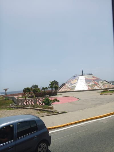 The image depicts a scenic view of a large, conical structure adorned with a colorful mosaic. The mosaic features figures and scenes, possibly historical or celebratory. The structure is situated in an outdoor setting with palm trees and other greenery surrounding it. A paved area with red accents and a low concrete wall with a decorative pattern is visible in the foreground, leading towards the structure. Beyond the greenery, a body of water, likely the ocean, can be seen, with waves gently rolling towards the shore. A distinctive sculpture, resembling a tree with a broad, flat top supported by a thick trunk, stands near the water's edge. In the lower left of the image, a blue car is partially visible, with a person seated inside. The scene appears to be a public park or monument area. The sky is clear and bright, suggesting it is daytime, likely late morning or early afternoon, with fair weather. The location cues, such as the palm trees and the proximity to the ocean, align with coastal regions. The overall mood is peaceful and open, with a sense of civic pride or historical commemoration conveyed by the monumental structure.
