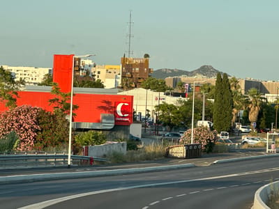 The image depicts an urban scene in La Valette-du-Var, France, likely a commercial area, characterized by modern architecture and a busy road. In the foreground, a multi-lane asphalt road curves into the frame, marked with white dashed lines. A concrete barrier separates the road from a sidewalk or pedestrian area. A guardrail is visible along the edge of this elevated area.
Dominating the mid-ground are several buildings. A large, bright red building with vertical corrugated siding is prominent on the left. Above its roofline, a smaller red structure with the white stylized letter "C" and the word "Charlemagne" is affixed. Adjacent to this red building is a white structure with large windows and a green awning. Further back, a brown, multi-story building with balconies and a distinctive antenna structure on its roof stands out against the sky. Other buildings in varying shades of white and yellow are scattered throughout the background.
Vegetation is interspersed throughout the scene, with lush green trees and a vibrant pink flowering bush on the left side of the red building. Palm trees are visible behind the buildings. The sky is a clear, pale blue, suggesting daytime with good weather.
In the background, a hilly, rocky landscape rises, typical of the Mediterranean region. Several cars are parked or driving in the visible areas, and some pedestrians can be seen near a bus stop shelter on the right side of the image. Trash receptacles are also visible. The overall impression is a snapshot of a functional urban environment, likely on the outskirts of a town or city, with elements of commerce, transportation, and residential or office buildings.