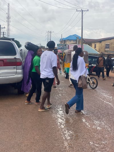 The image shows a street scene in Barakin, Nigeria, with several people and objects. A group of individuals are standing around a silver car, some appearing to be engaged in conversation or preparing to load items into the vehicle. One person in the foreground is wearing a green t-shirt and black pants, with a purple cloth draped over them and the car's trunk. Another person in a white t-shirt and black shorts is standing next to them, looking towards the side.
Further in the background, a woman in a white shirt and blue jeans is walking, carrying a brown bag. Next to her, a small motorized tricycle, common in Nigeria, is visible. Other people are also seen in the distance, walking along the street or near buildings. The setting appears to be a moderately busy urban area with shops and buildings lining the street. Utility poles and power lines are prominent in the background, suggesting an established infrastructure. The ground is paved with asphalt and shows signs of recent rain or cleaning, with white splotches scattered across the surface. The sky is overcast, indicating a cloudy or possibly humid day. The overall atmosphere seems to be one of everyday activity and movement within a local community. The text "AMEW" is visible on the back of the white t-shirt in the foreground.