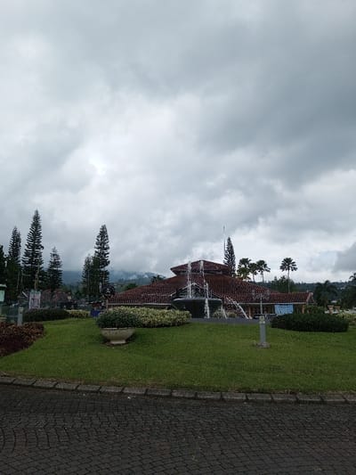 A fountain stands in front of a building with a red-tiled roof and lush, green lawn. Evergreen trees dot the landscape behind the building, while palm trees peek out from the right. The sky is overcast with heavy, gray clouds obscuring the sun. A paved brick road stretches across the lower part of the frame. Potted plants and manicured flower beds add to the scene's overall manicured appearance.