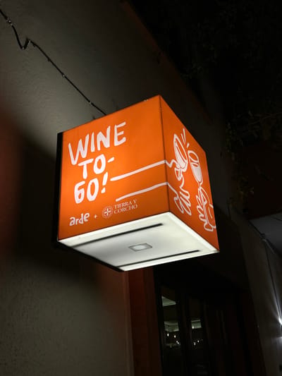 A brightly lit, orange, cube-shaped sign hanging on the wall of a building. The sign reads "Wine To-Go!" in white lettering. Above that are white line drawings of two glasses, one filled with liquid, and two hands reaching up. The other visible side of the cube sign is white with "arde + TIERRA Y CORCHO" printed in smaller letters. The wall is a rough, light gray texture with some electrical wires running up it. Above the sign, a dark window is visible. The sign appears to be advertising a wine service in a location, and based on the brand, is likely Mazatlán, Mexico. The scene is dimly lit, implying it is nighttime.