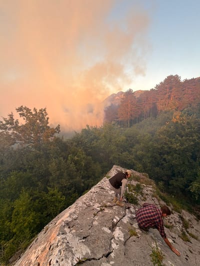 In Kanjoši, Montenegro, two individuals are positioned on a rocky, uneven terrain overlooking a dense forest. Smoke, tinged with orange hues from what appears to be a distant fire, drifts across the sky, obscuring the upper portion of a forested mountainside. The foreground is dominated by the rough, gray rock face, with sparse vegetation growing in its crevices. One person, wearing a dark vest over a lighter shirt and gray pants, is crouched down, holding a metallic water bottle. The other person, dressed in a red and black plaid shirt and gray pants, is further down the slope, also crouched and reaching out with their right hand. The trees in the mid-ground are lush and green, contrasting with the smoky sky and the arid rock. The lighting suggests it may be late afternoon or early evening, given the warm tones of the smoke and the sky.