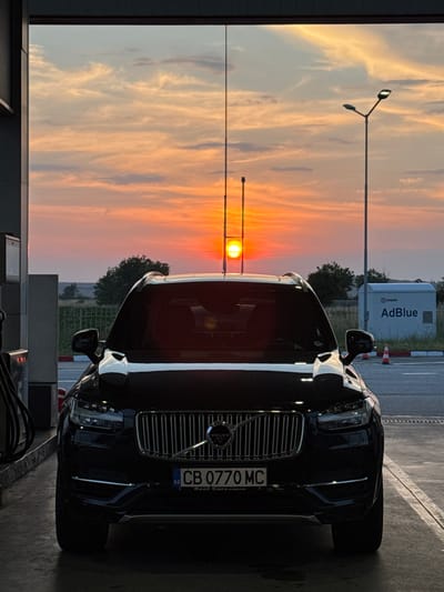 A black Volvo XC90 SUV is depicted head-on, parked under a fuel station canopy in Belozem, Bulgaria, during a vivid sunset. The vehicle occupies the lower center of the frame, its dark paint reflecting ambient light while the foreground remains underexposed against the bright sky. The front grille, prominently featuring the Volvo emblem, and the distinctive "Thor's Hammer" LED headlights are visible. The Bulgarian license plate reads "CB 0770 MC" with "BG" and "Real Cars 24.mobile.bg" text below. To the left, a grey fuel pump and black hoses are partially visible.
The background is dominated by a dramatic sunset sky, showcasing a palette of intense orange, red, and yellow hues near the horizon, transitioning to lighter orange and blue tones higher up. The bright, circular sun is centered directly behind the car, partially obscured by the vehicle's roof antenna. Scattered clouds with defined textures are present across the sky.
On the right side of the frame, a street light with an illuminated LED fixture stands prominently. To its left, another pole is visible. Further right and behind the car, a white rectangular container bearing the labels "ЕКОНОЙЛ" and "AdBlue" is situated beside an asphalt road. A red and white traffic cone is placed on the road near the container. Beyond the immediate fuel station area, a wire fence separates the road from a distant, flat landscape featuring scattered green trees and what appears to be open agricultural land under the expansive sky. No people are present in the scene.