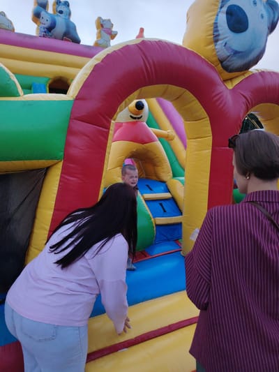 In the city of Олтушскі сельскі Савет, Belarus, a child is crying while on an inflatable castle. The castle is brightly colored with red, yellow, green, and pink sections, featuring cartoon characters at the top. Two women are observing the child from outside the castle. One woman, with long dark hair, wears a light pink long-sleeved shirt and light blue jeans. The other woman, with short brown hair, is wearing a maroon and pink striped shirt and sunglasses on her head. The environment appears to be outdoors, with a cloudy sky visible in the background.