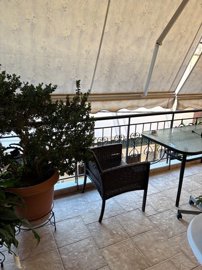 The image shows an outdoor seating area, likely a patio or balcony, with a wicker chair and a glass-topped table. The setting appears to be a residential or commercial space, possibly a cafe or restaurant, given the presence of outdoor furniture. A large potted plant with dark green leaves is prominent in the foreground, partially obscuring the view of the furniture. A white peace lily is also visible in the pot, with its distinctive white spathe.
The area is covered by an awning made of light-colored fabric with a floral pattern. The awning is supported by a metal frame, with one section extended over the seating area. The ground is tiled with large, light brown square tiles. In the background, a black metal railing surrounds the area, and beyond that, a building is visible with windows.
The time of day appears to be daytime, with natural light illuminating the scene. The weather seems pleasant, suitable for outdoor dining or relaxation. There are no people visible in the image. A small white label with the number "28" is visible on a surface in the background, possibly a door or wall. The overall impression is one of a tranquil and inviting outdoor space.