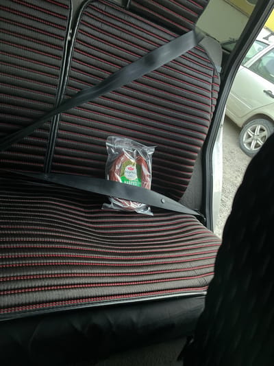 In Buynaksk, Russia, a plastic-wrapped package of sausage is resting on the black and red striped fabric seat of a car's backseat. A black seatbelt diagonally crosses the package and the seat. The car appears to be parked outdoors, as evidenced by the asphalt and another vehicle visible in the background.