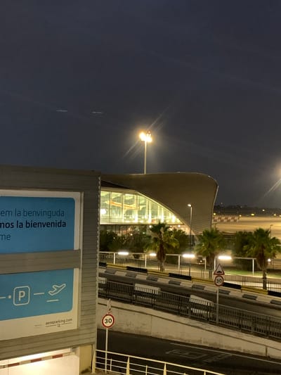 This is a vertical, nighttime photograph taken in Manises, Spain. The scene captures a modern building with a curved roof and a glass facade, illuminated from within, suggesting it's likely an airport terminal or related facility. Several palm trees are visible, indicating a warm climate.
In the foreground, a blue sign features white text and icons. Part of the text reads "em la benvinguda" and "mos la bienvenida", which are Catalan and Spanish phrases for "welcome". An icon of an airplane and a "P" in a square, likely indicating parking, are also present. The website "aenaparking.com" is visible at the bottom right of the sign.
Further in the background, a road or pathway runs alongside the building, bordered by metal railings. A speed limit sign indicating "30" is mounted on a pole, suggesting a controlled speed environment. Streetlights cast a warm glow on the scene, and the sky is dark, indicating it is nighttime. The overall impression is one of arrival or departure at a well-lit, modern facility.