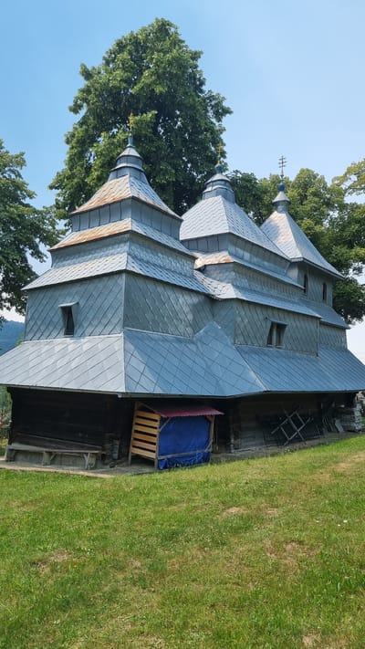 The image depicts a wooden church with multiple tiered roofs covered in grey metal shingles. The church has a dark wooden lower section. In front of the church, there is a simple wooden bench on the left and a small wooden structure with a red roof covered by a blue tarp on the right. The ground in front of the church is covered in green grass. Behind the church, there are tall green trees and a clear blue sky. The overall scene suggests a rural setting, likely in Vyshka, Ukraine, given the architectural style often found in that region. There are no people visible in the image.