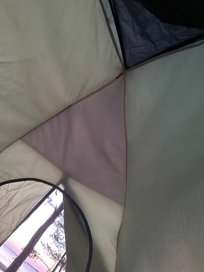 The image presents a view from inside a tent, looking outwards towards a natural landscape during twilight hours, likely sunrise or sunset. The interior of the tent shows a mix of light cream-colored and darker gray fabric panels, suggesting the inner lining and possibly an outer shell or partition. A small red loop or tie is visible near the seam of two fabric sections.
Through an open, circular tent flap or entrance, a serene outdoor scene unfolds. Dark, silhouetted trees, appearing to be pines, stand against a colorful sky. Beyond the trees, a calm body of water, possibly a lake or large river, reflects the soft light of the sky. The sky itself displays gentle hues of pink, purple, and orange, characteristic of dawn or dusk.
The overall setting is one of peaceful camping in nature. No people are visible, emphasizing the tranquility of the moment. The location is Zerendi, Kazakhstan, implying a scenic natural area in that region. The image conveys a sense of calm and a quiet observation of the natural world from the comfort of a tent. There is no visible text in the image.