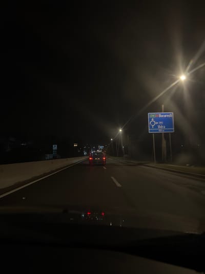 The image depicts a nighttime scene on a multi-lane asphalt road in Jilava, Romania, likely on the E70 route approaching the turn-off for Vidra and Bucharest. The perspective is from inside a vehicle, with the dashboard visible at the bottom.
Ahead, a grey sedan with illuminated red taillights is visible in the same lane, followed by additional vehicle lights in the distance, indicating traffic. To the right, a prominent blue road sign displays "E70 București" (Bucharest) at the top, and below it, a roundabout symbol with "Vidra 150m," indicating an upcoming turn towards Vidra via a roundabout in 150 meters.
The road surface is dark, with reflective patches possibly from moisture. White lane markings are visible: a solid line bordering the left side of the current lane and a dashed line separating lanes further ahead. A light-colored concrete barrier runs along the left side of the road. Multiple streetlights illuminate the scene, with one on the right creating a prominent starburst lens flare. Another streetlight is visible further down the road on the left. A smaller, lighter-colored sign is partially visible beyond the concrete barrier on the left. The sky is dark and devoid of visible stars.