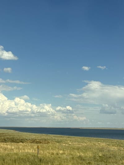 The image depicts a wide, panoramic view of a rural landscape under a bright, partly cloudy sky. The scene is oriented vertically, with the ground occupying the right side and the sky the left. The ground is a gently sloping field of dry, yellow-green grass, suggesting a warm season or dry climate. A dark, linear feature runs horizontally through the middle ground, likely a body of water or a distant treeline.

In the foreground of the grassy field, a small, yellow object resembling a post or marker stands upright. The sky is a vibrant blue, dotted with fluffy white cumulus clouds scattered across the expanse. The clouds are more concentrated towards the center and right of the frame, with some appearing wispy and others more voluminous.

The lighting suggests it is daytime, with the sun high in the sky. The overall atmosphere is one of natural serenity and open space, indicative of a rural or wilderness setting. There is no text visible in the image. The location context provided places this scene in Divnoe, Russia, which is consistent with an open, grassland environment.