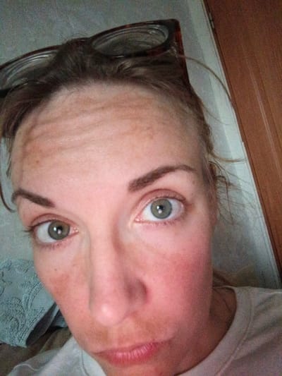 The image is a close-up, eye-level selfie of a woman's face, taken indoors. The woman has light skin with freckles and visible forehead wrinkles. Her hair is dark brown and pulled back, with some strands loose around her face. She is wearing glasses with dark frames, perched on her head. Her eyes are open and appear to be light green or grey. Her expression is neutral, and her mouth is slightly open, revealing her teeth. The background is blurred and consists of a textured light-colored wall and a wooden door on the right side. A piece of textured, light blue fabric is visible on the left side of the frame. There is no indication of the city of Malaya Vishera, Russia, in the image or its context.