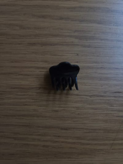 A small, black hair claw is positioned centrally on a wooden surface. The hair claw is made of plastic and has four prongs that extend outwards. The wooden surface is made up of vertical planks with a visible grain pattern. The lighting suggests it is daytime, with a shadow cast by the hair claw to the left. There is no text visible in the image, and no people or animals are present. The setting appears to be indoors, given the artificial lighting and the nature of the surface. The image is a close-up shot, focusing solely on the hair claw and the wooden background.